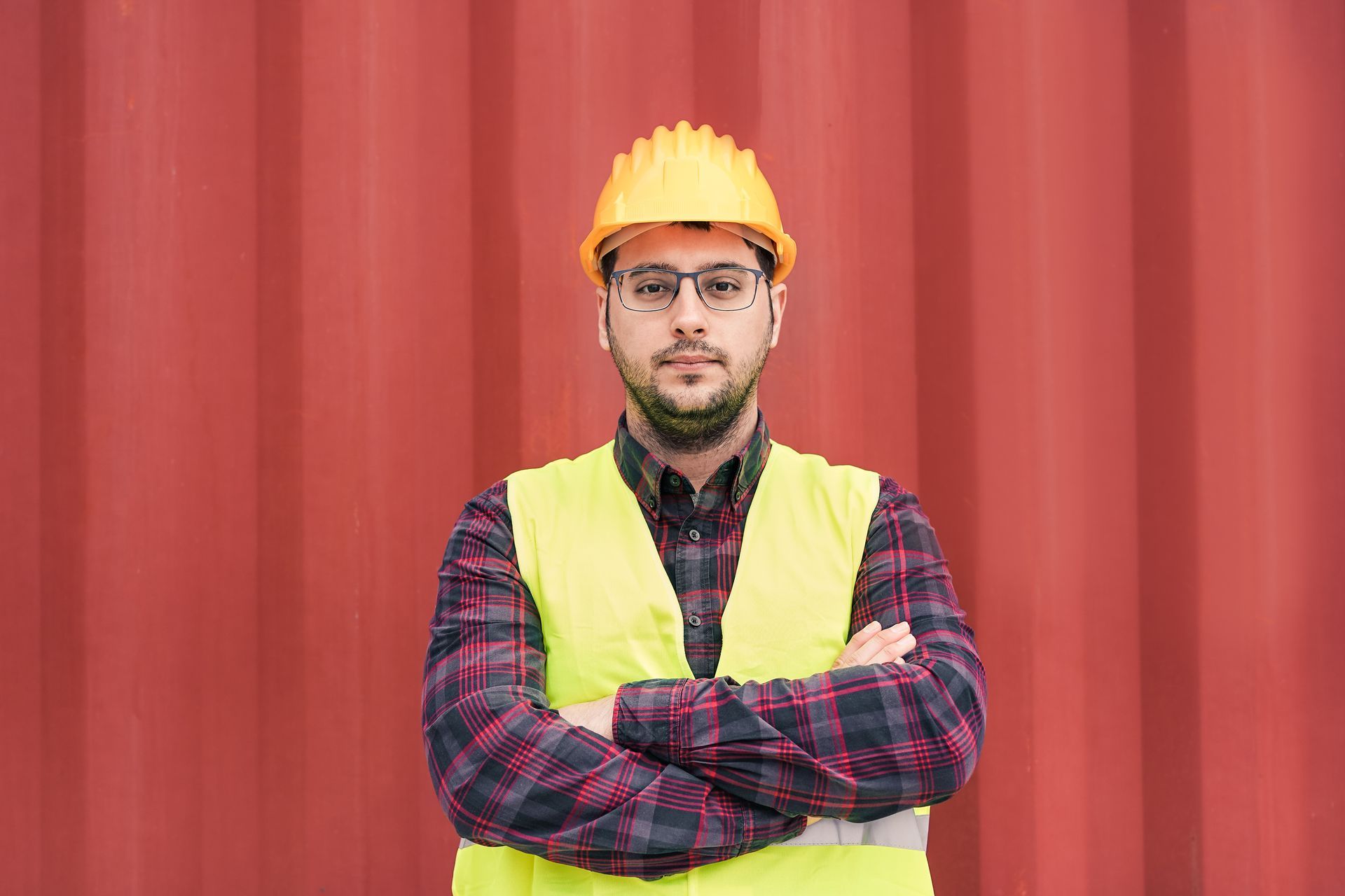Man in yellow hard hat and vest, arms crossed, in front of a red wall.