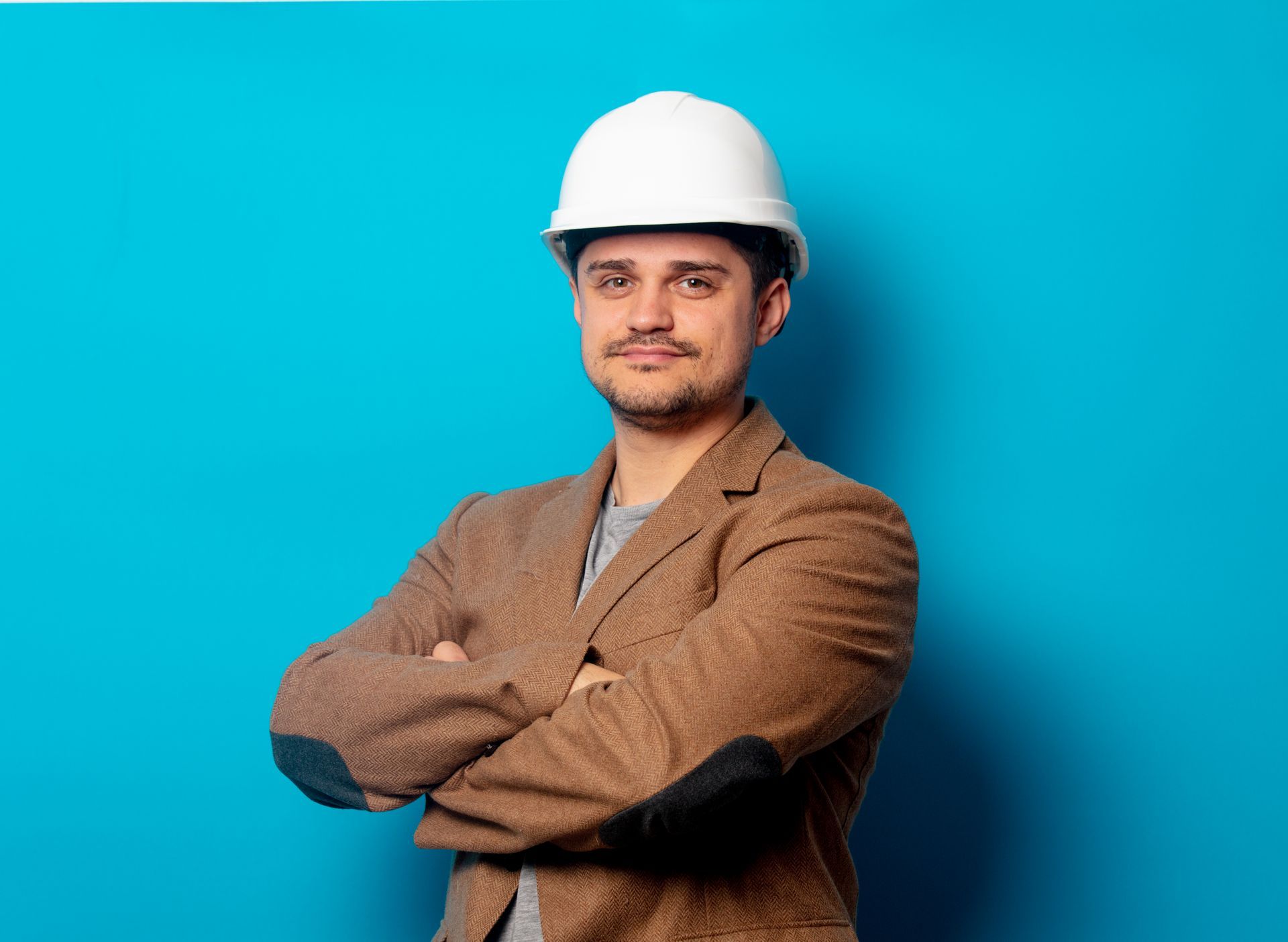 Man in white hard hat and brown jacket, arms crossed, smiling against a blue background.