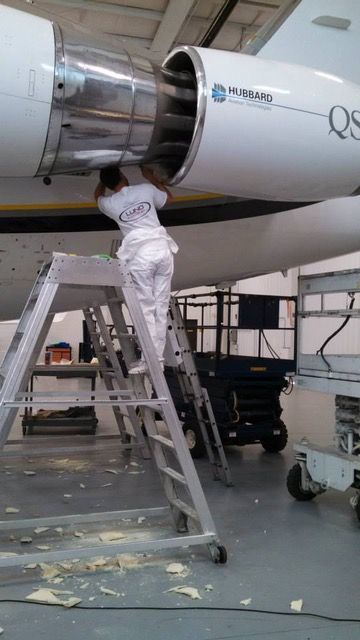 Person working on an aircraft engine using a ladder in a hangar.