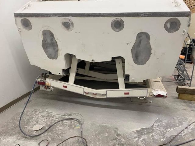 A boat on a trailer, rear view, in a workshop. Boat is white with gray patches, trailer is also white.