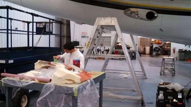 Person working on materials at a table under an airplane, using ladder.