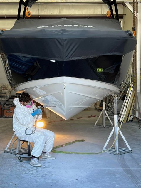 Person in protective suit spraying a white boat's hull, supported by stands, in a workshop.