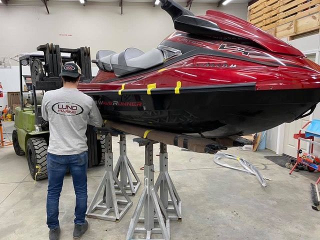 Man near a red jet ski lifted by a forklift, supported by jack stands, in a workshop.