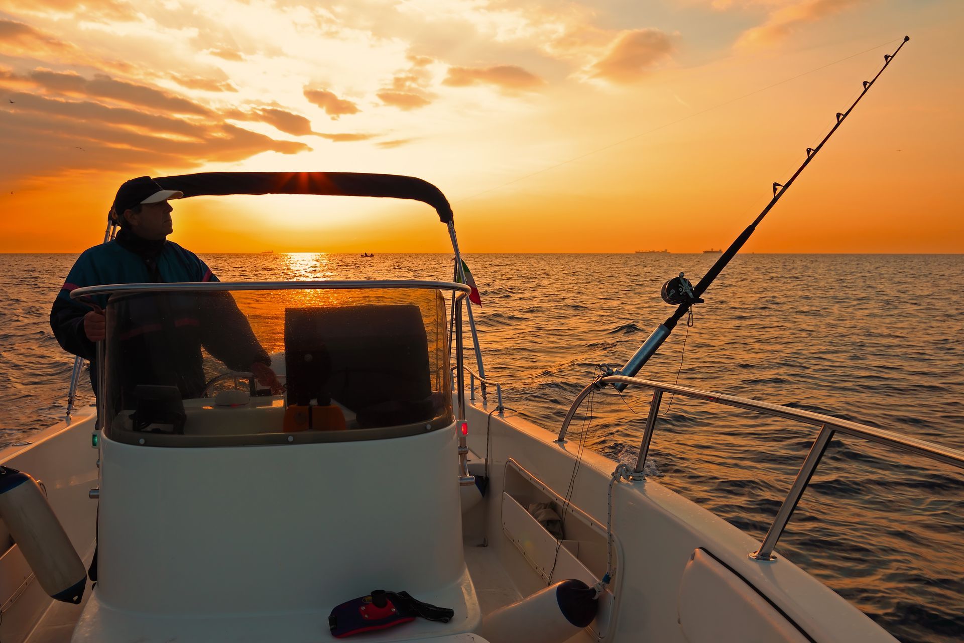 Man piloting a fishing boat at sunset with fishing rod, orange sky.
