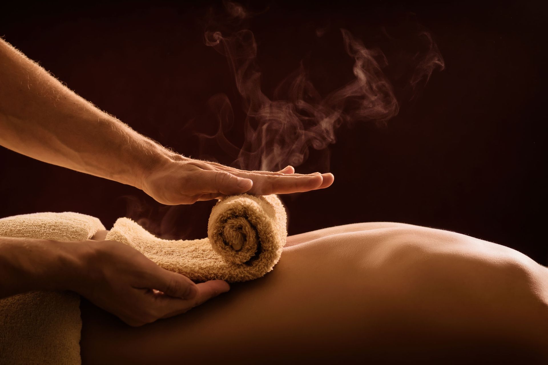 Hands giving a hot stone massage to a person’s back, with steam and warm brown tones