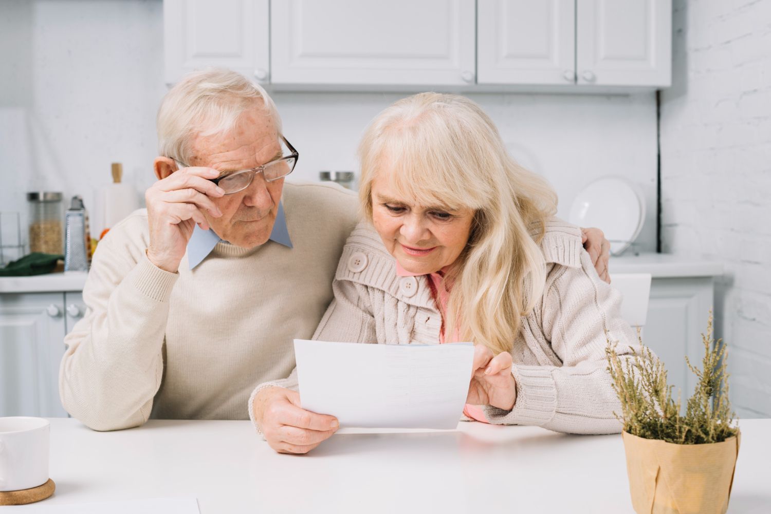 Elderly couple reviewing document at a table. Man adjusting glasses, woman smiling, both in a kitchen.