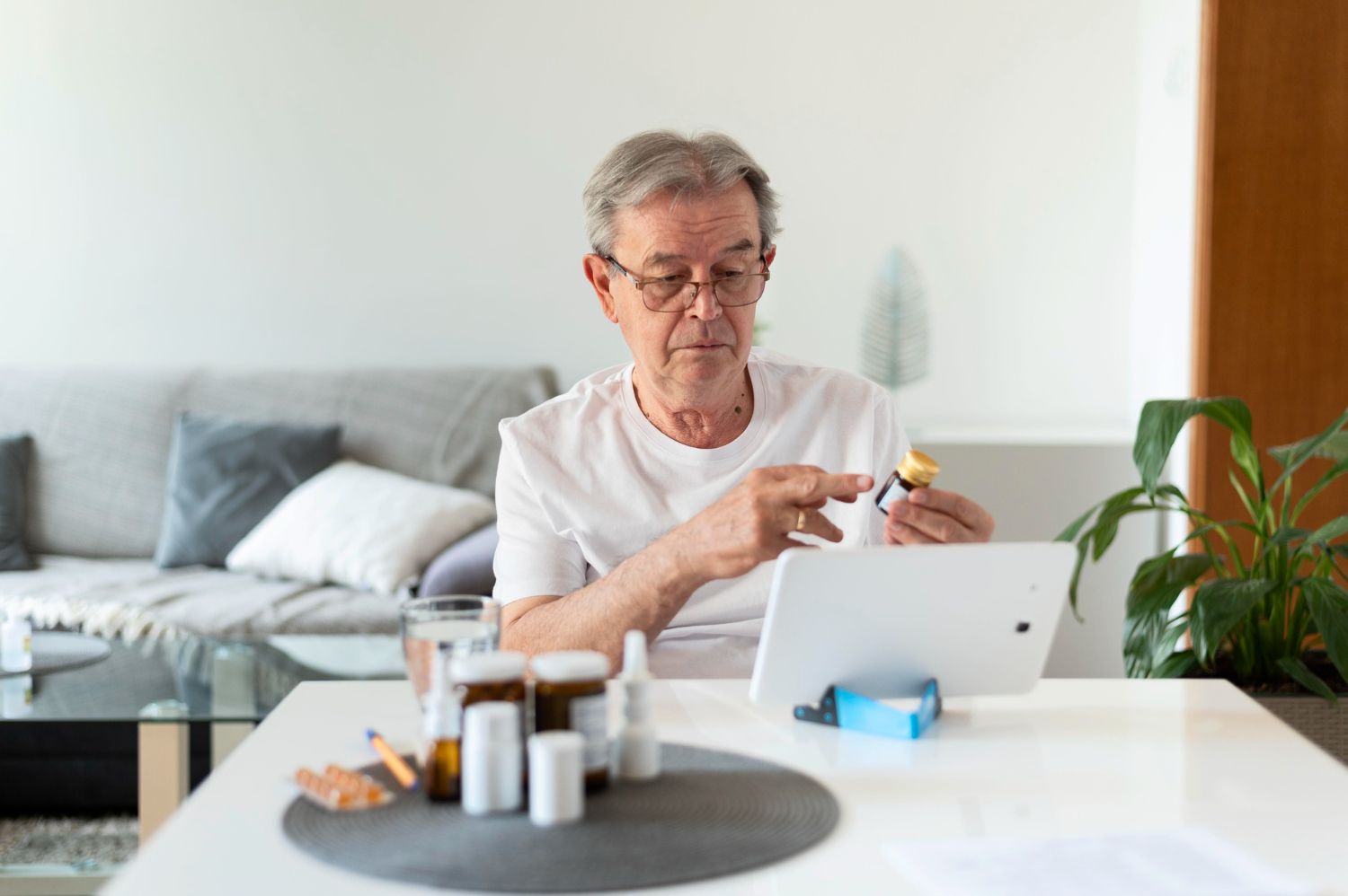 Older man at table, video call, holding pill bottle, tablet, other medications nearby.