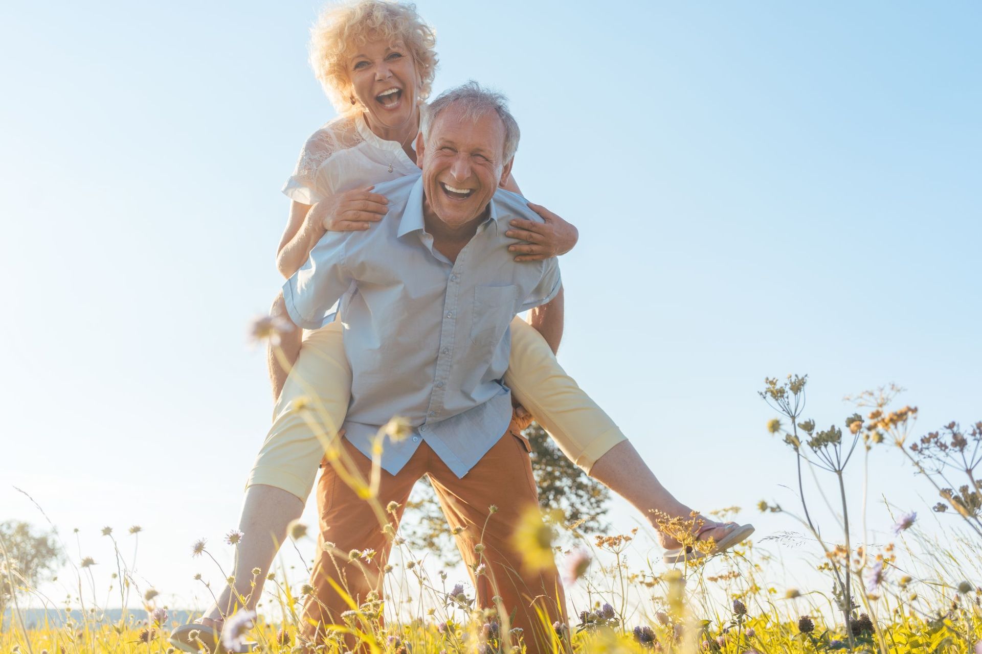 Smiling couple in a field; man giving woman a piggyback ride; blue sky and sunlight.