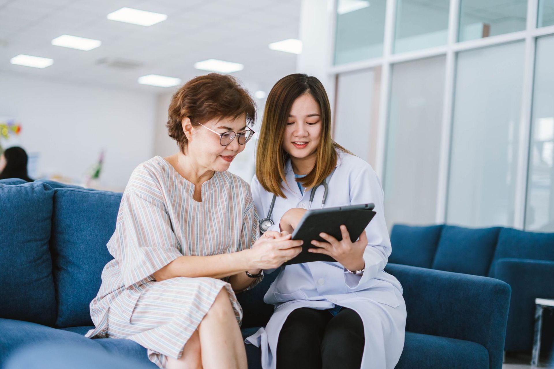 Doctor showing tablet to patient, both smiling on blue sofa in hospital.