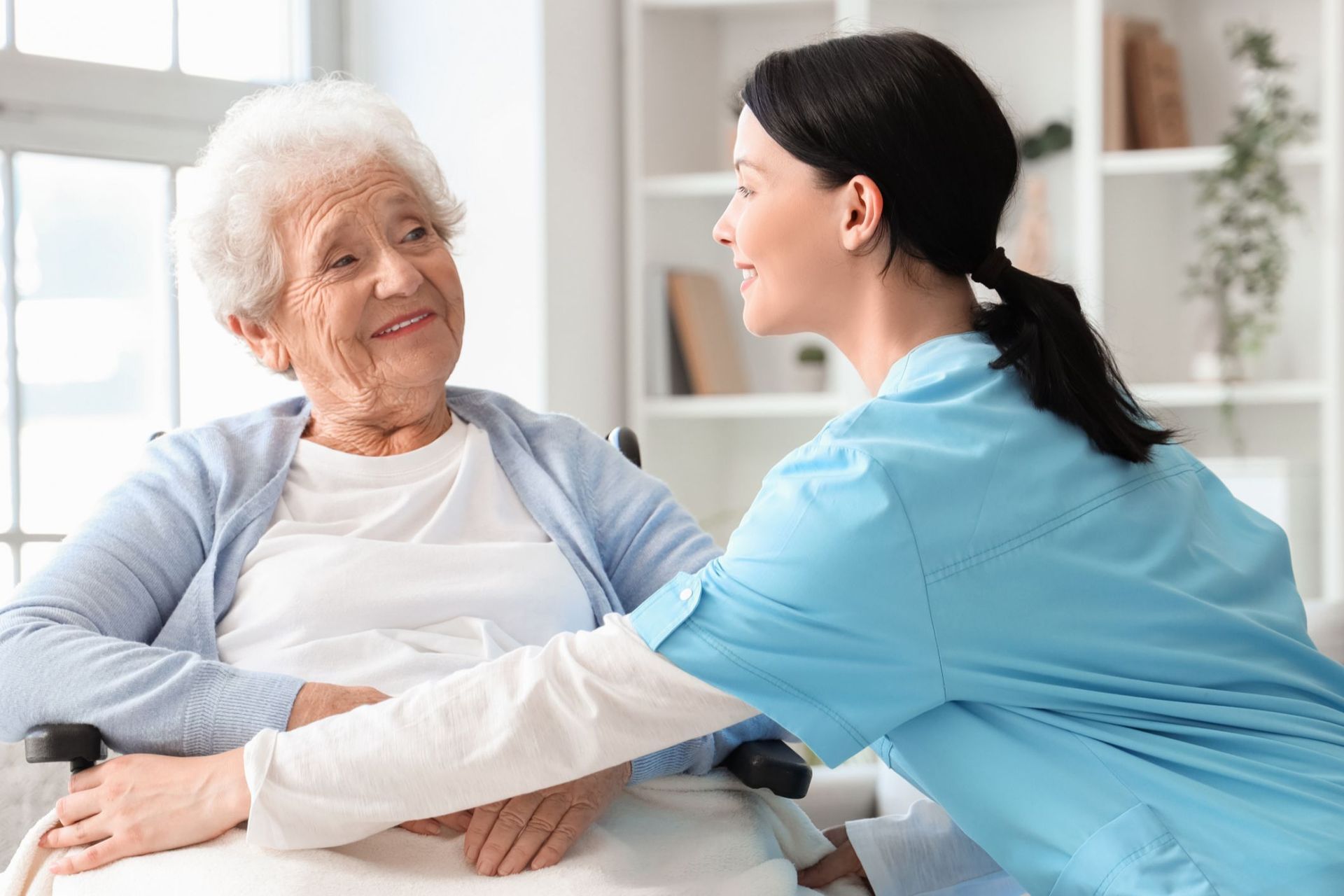 A caregiver in blue scrubs assists an elderly woman in a wheelchair; both are smiling.