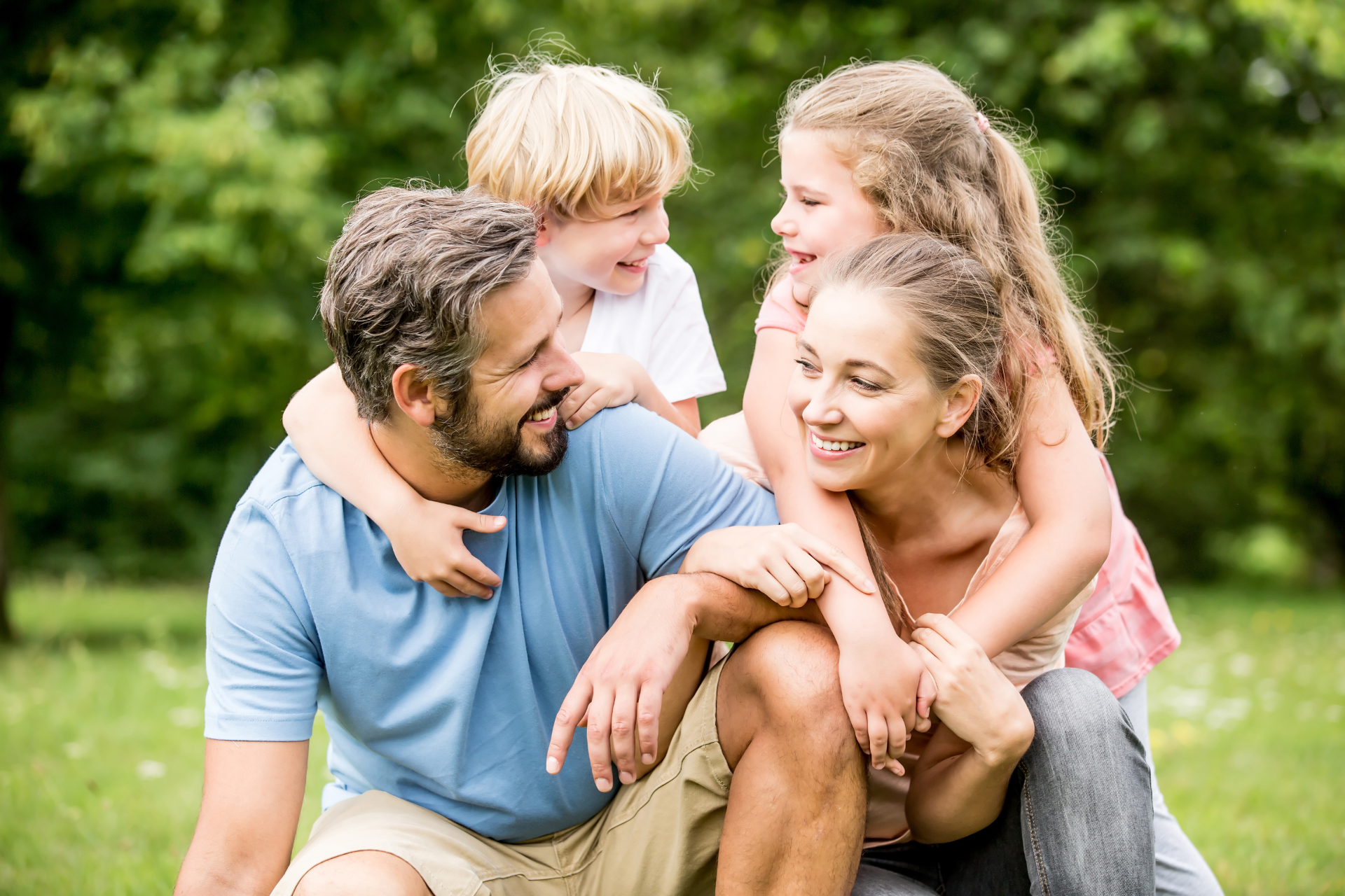 Family of four embracing outdoors; parents smiling, children laughing.