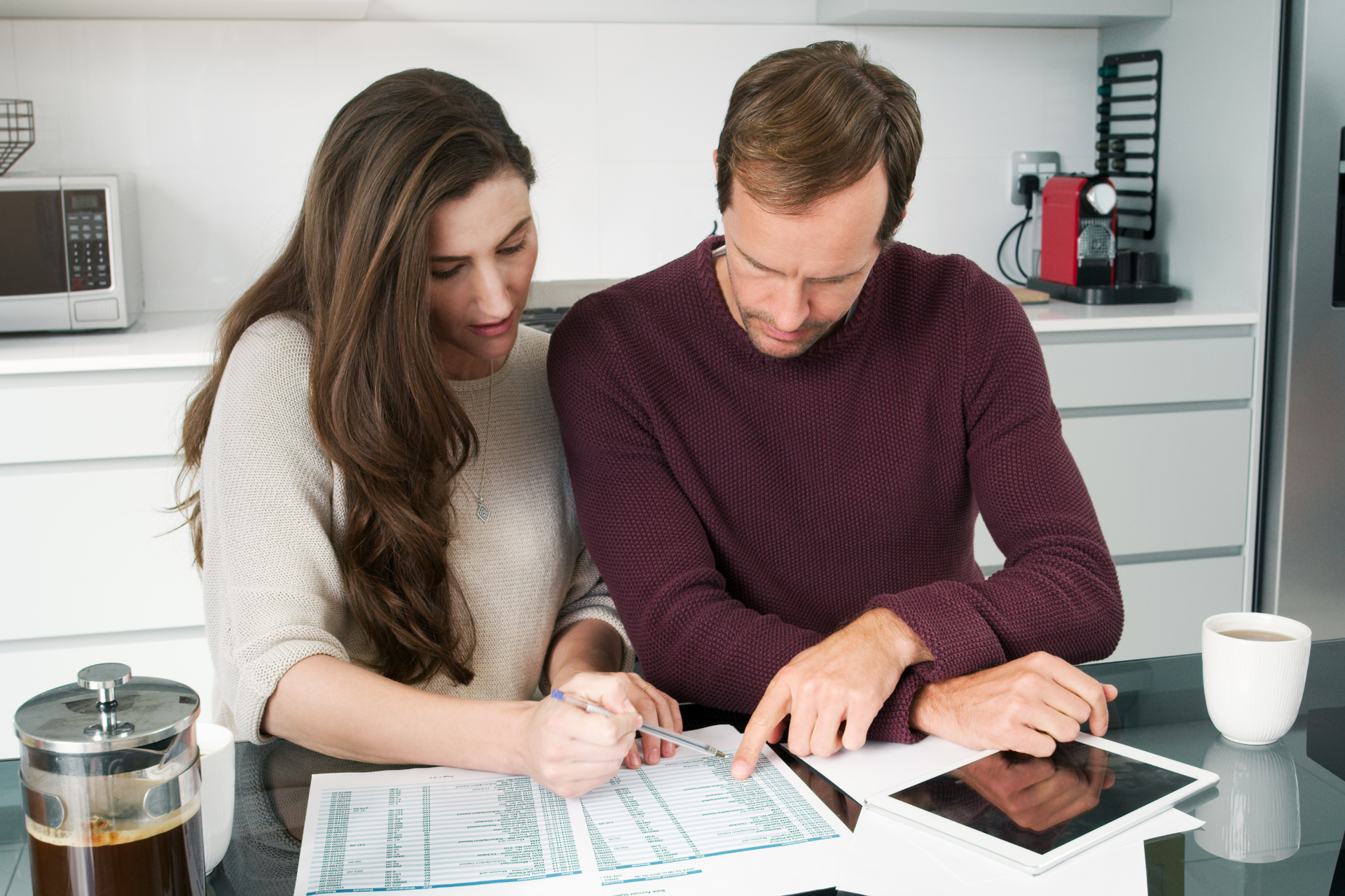 Couple reviewing papers at a kitchen counter, coffee and tablet present.