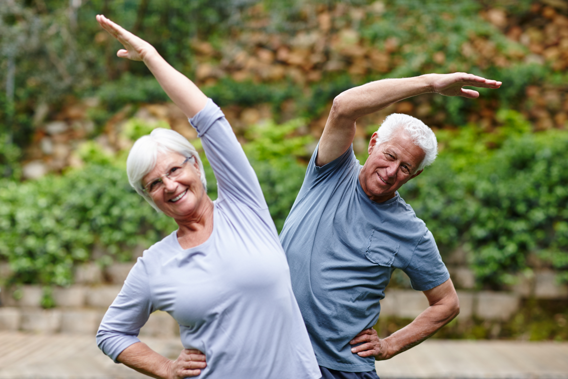 An older couple exercises outdoors, smiling and stretching their arms.