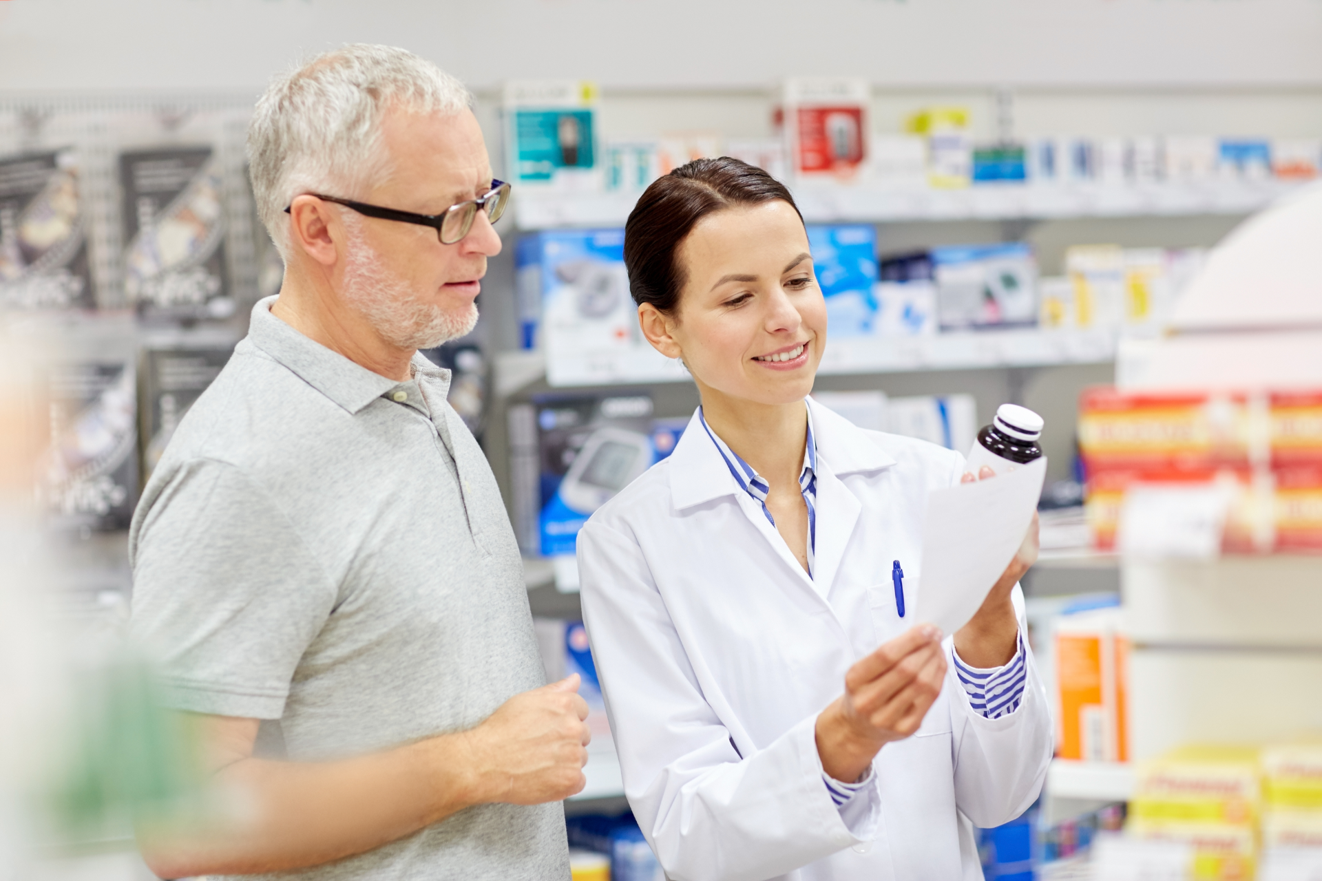 Pharmacist in white coat explains medication label to older man in a pharmacy.