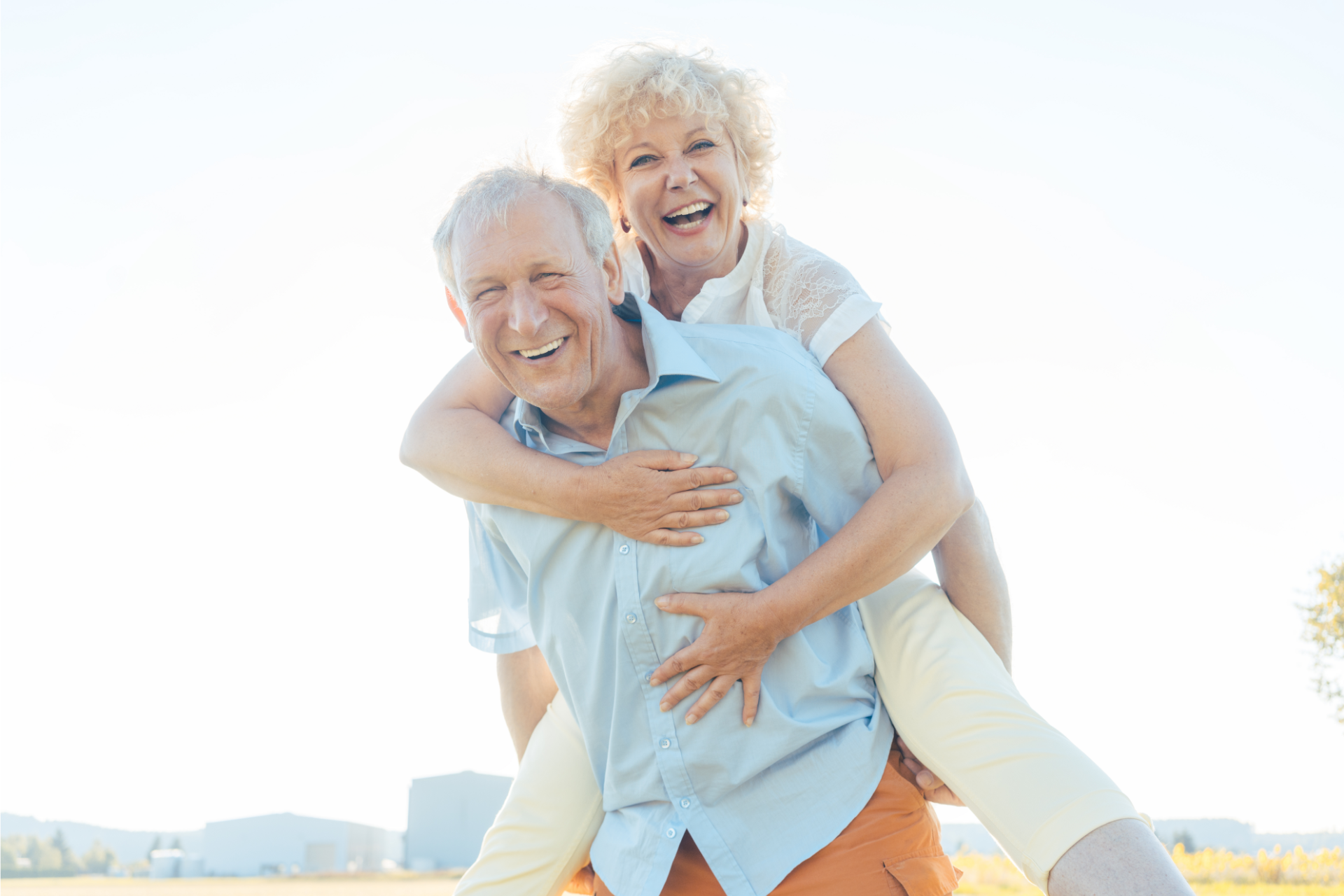 Elderly man giving a piggyback ride to smiling woman in sunny field.