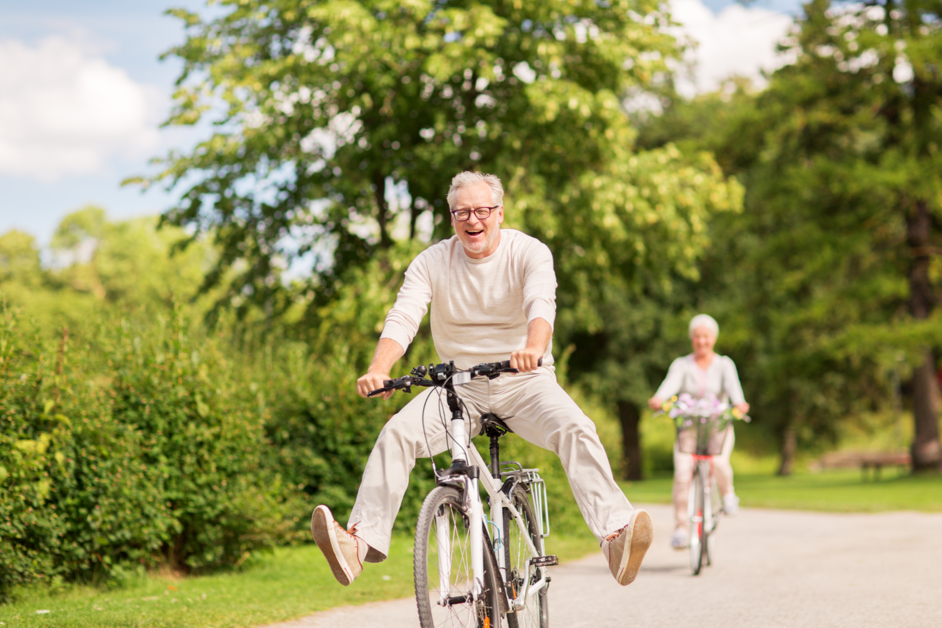 Happy elderly man riding bicycle, arms outstretched, in park with woman cycling behind.