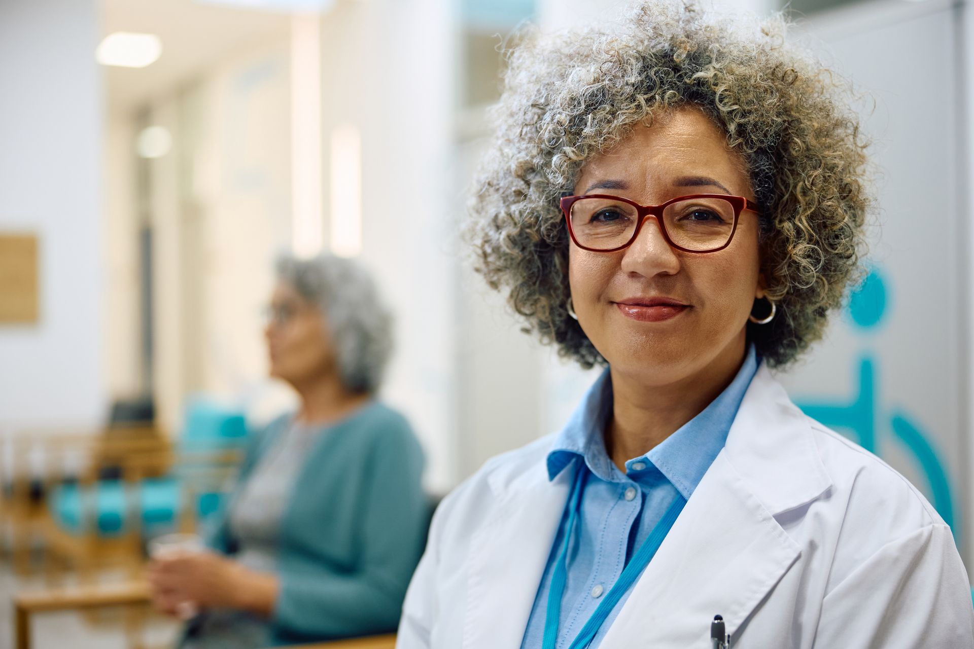 Female doctor with curly gray hair smiles at the camera, a patient seated behind her in a waiting room.