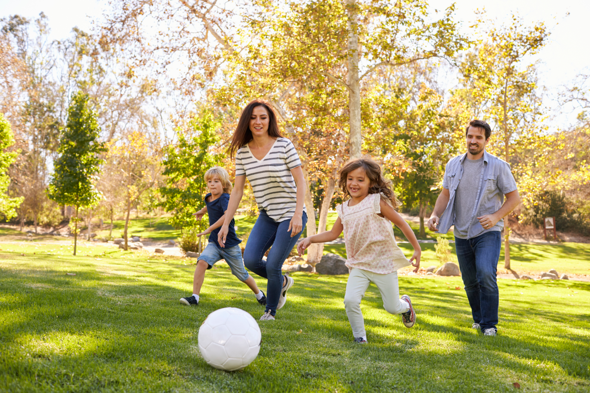 Family of four playing soccer in a sunny park; they are laughing and running after the ball.