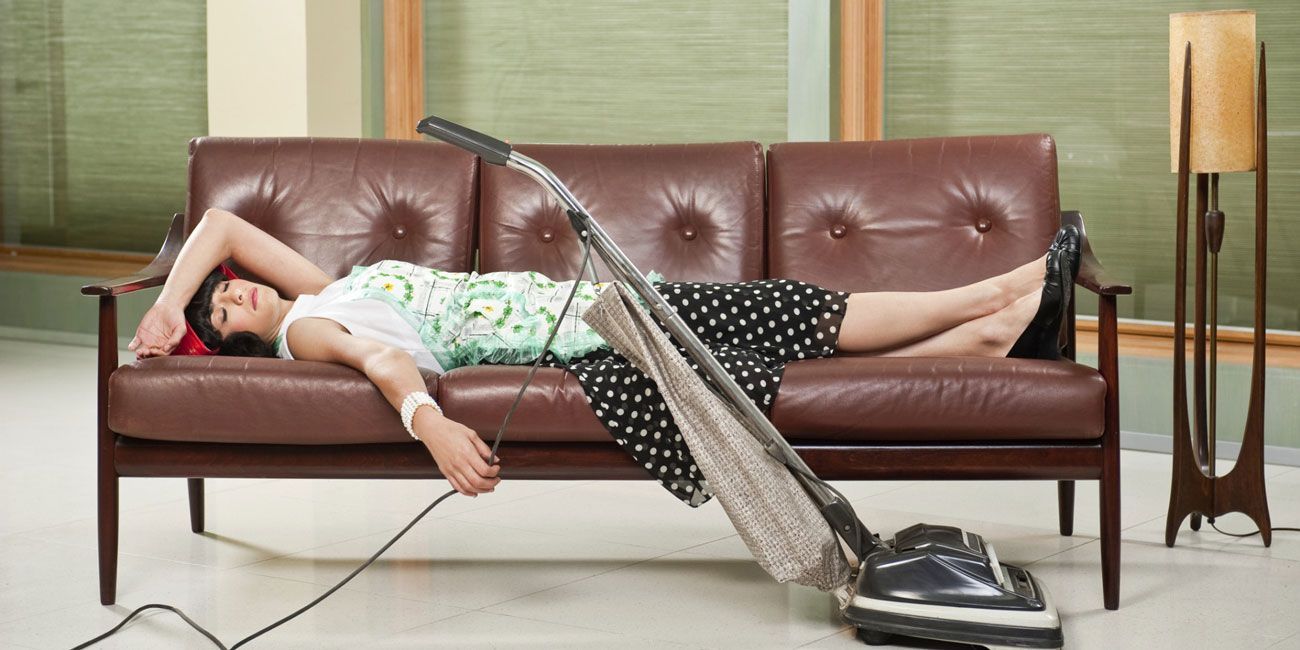 A woman is laying on a couch next to a vacuum cleaner.