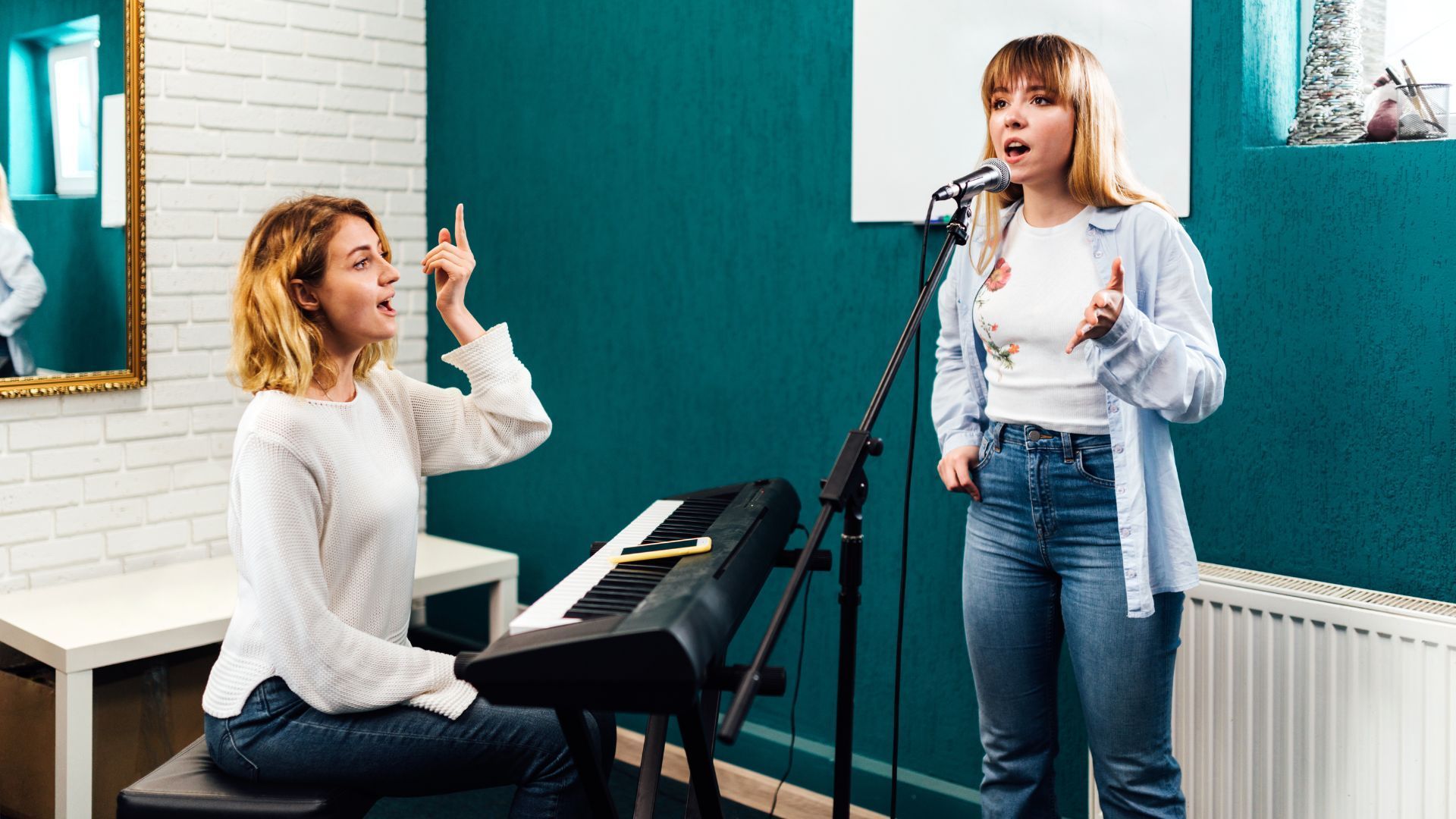 Woman singing into a microphone, with another woman at a keyboard gesturing. Indoor studio setting.