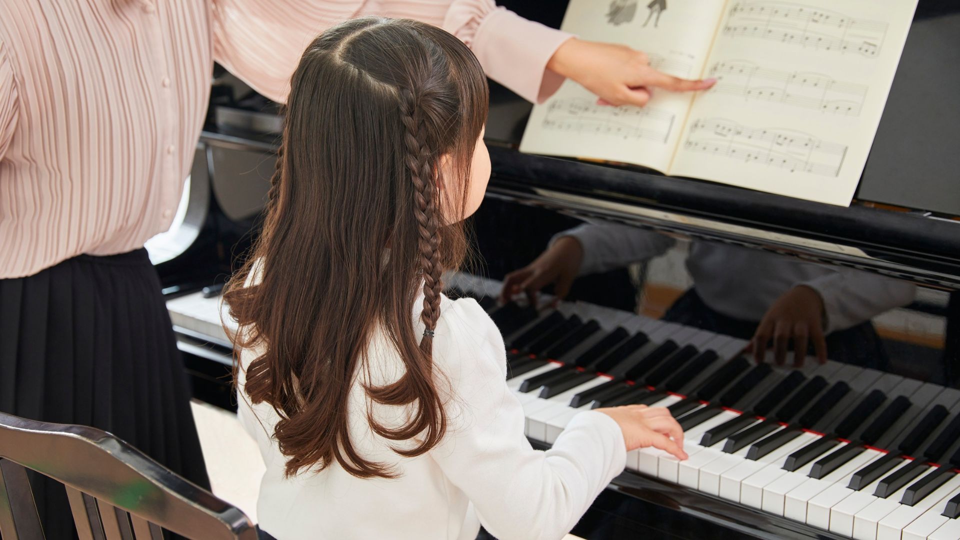 Young child playing piano with instructor pointing at sheet music.