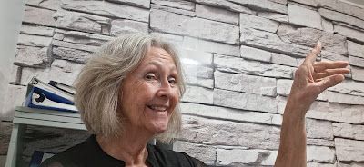 Woman making a hand gesture, smiling, against a textured gray brick wall background.
