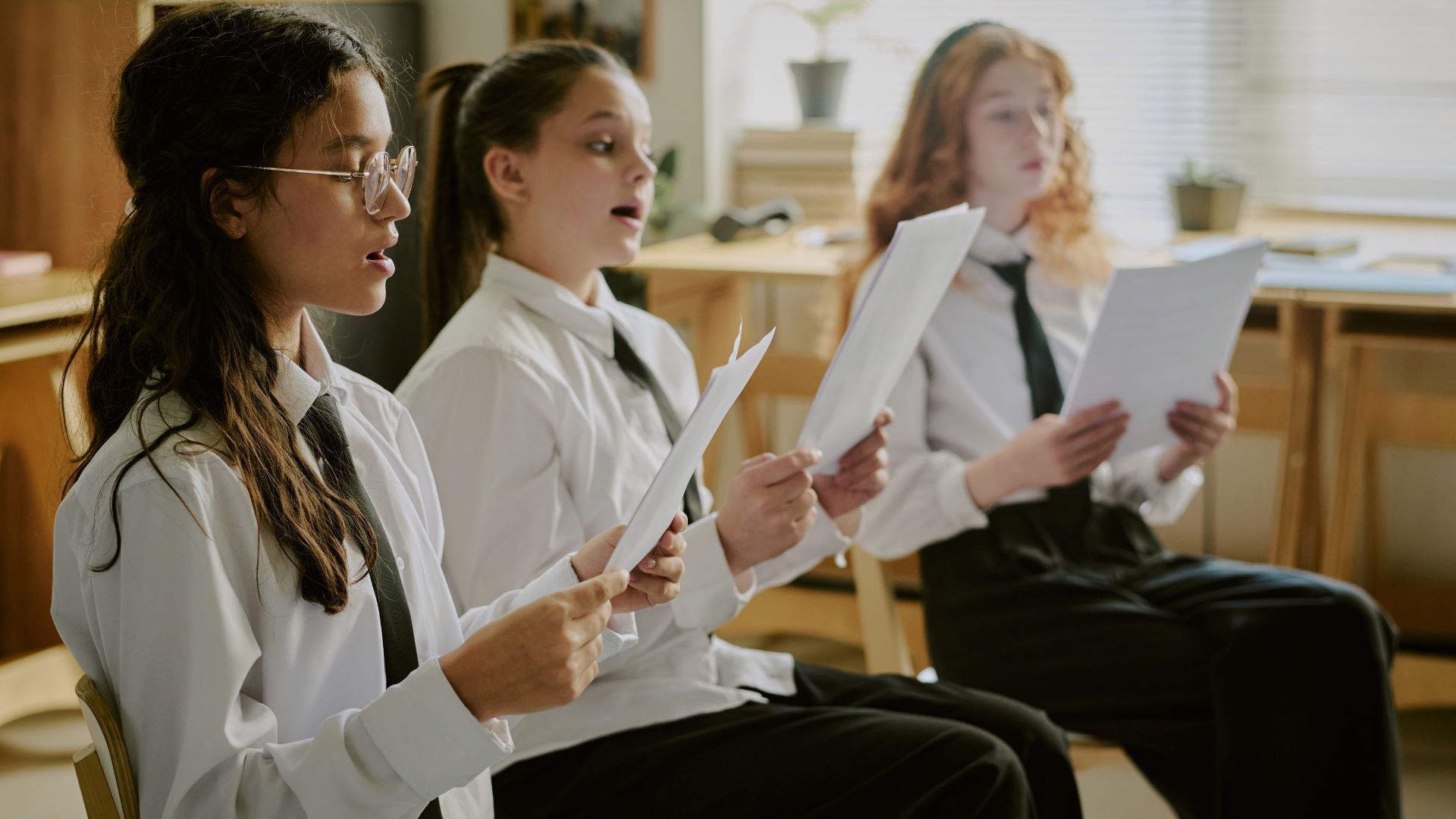 Three girls in white shirts and ties sing, holding sheet music.