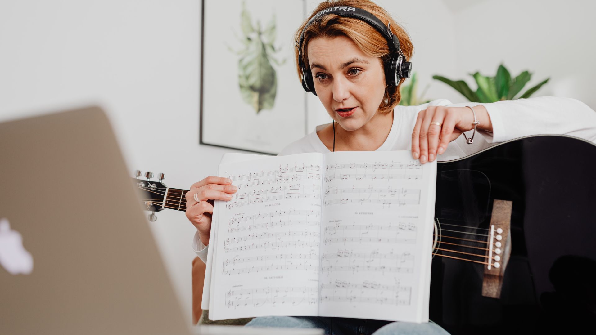 Woman with headphones teaching guitar online, holding sheet music, laptop visible.