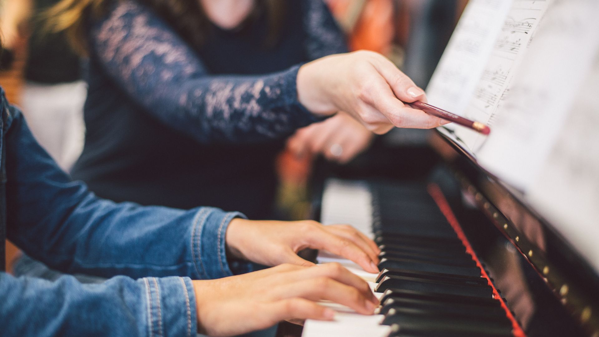 Person's hands playing piano, another person's hand pointing at sheet music. Indoor setting.