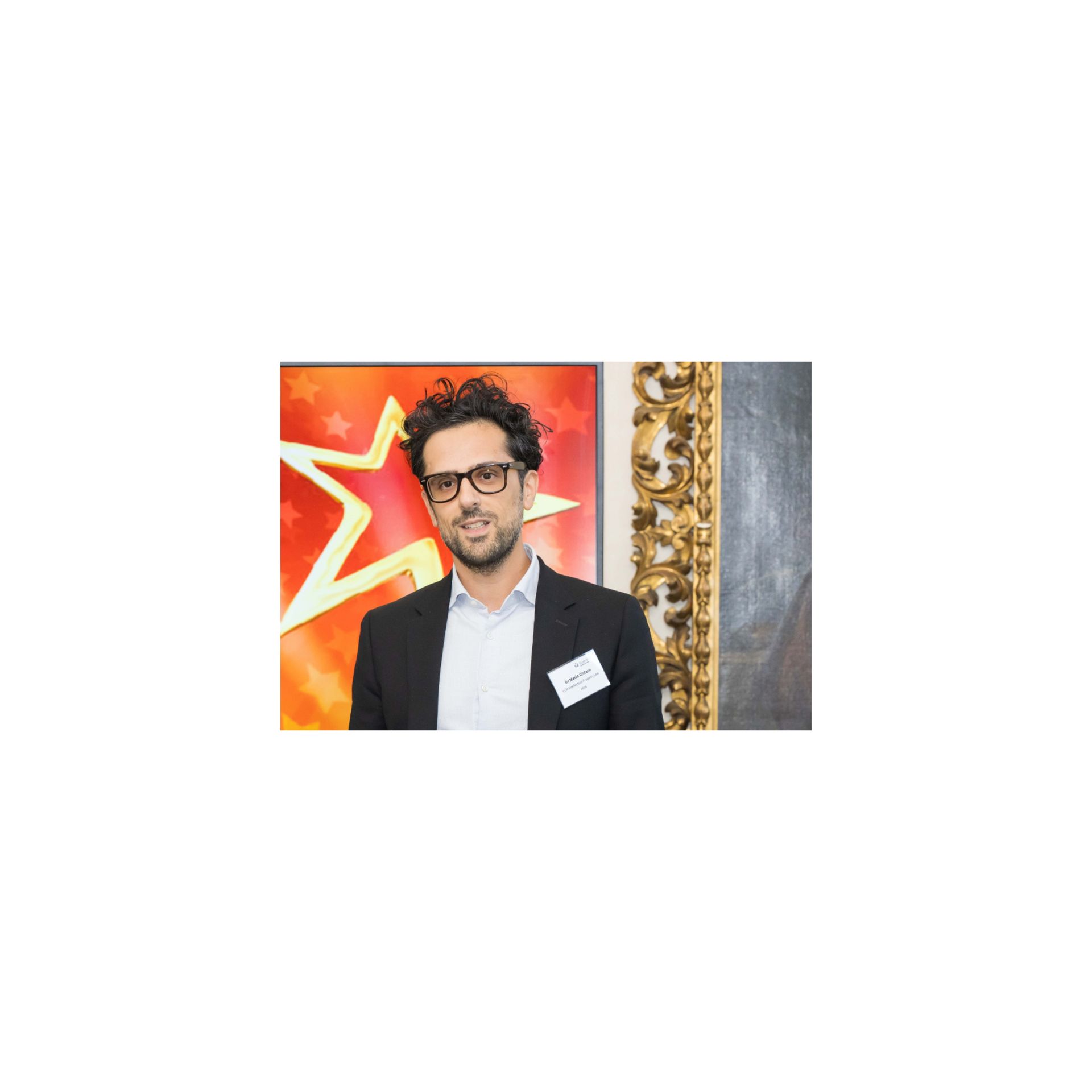 Man with curly hair and glasses in a black suit, standing indoors near a gold decorated wall.