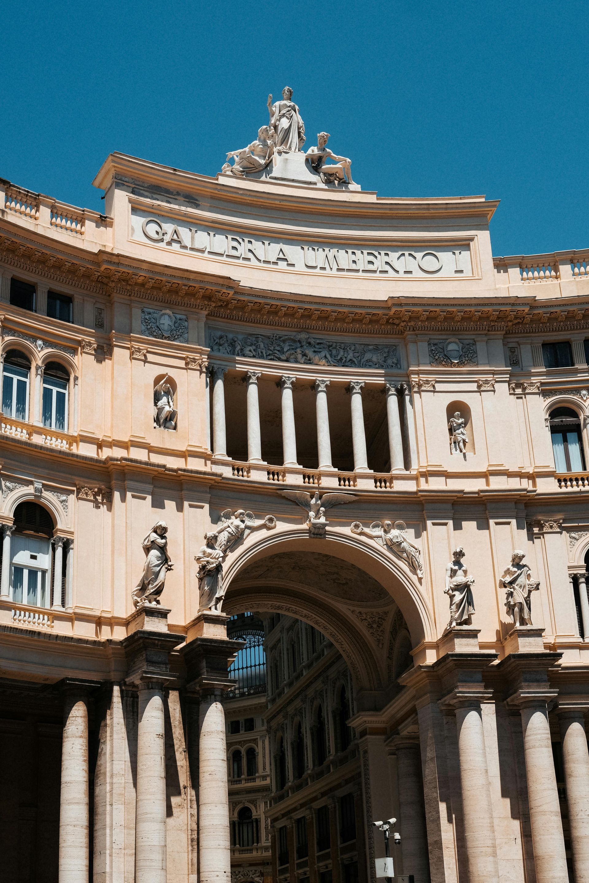 An arched, ornate building facade in Naples, Italy. Cream-colored with a blue sky.