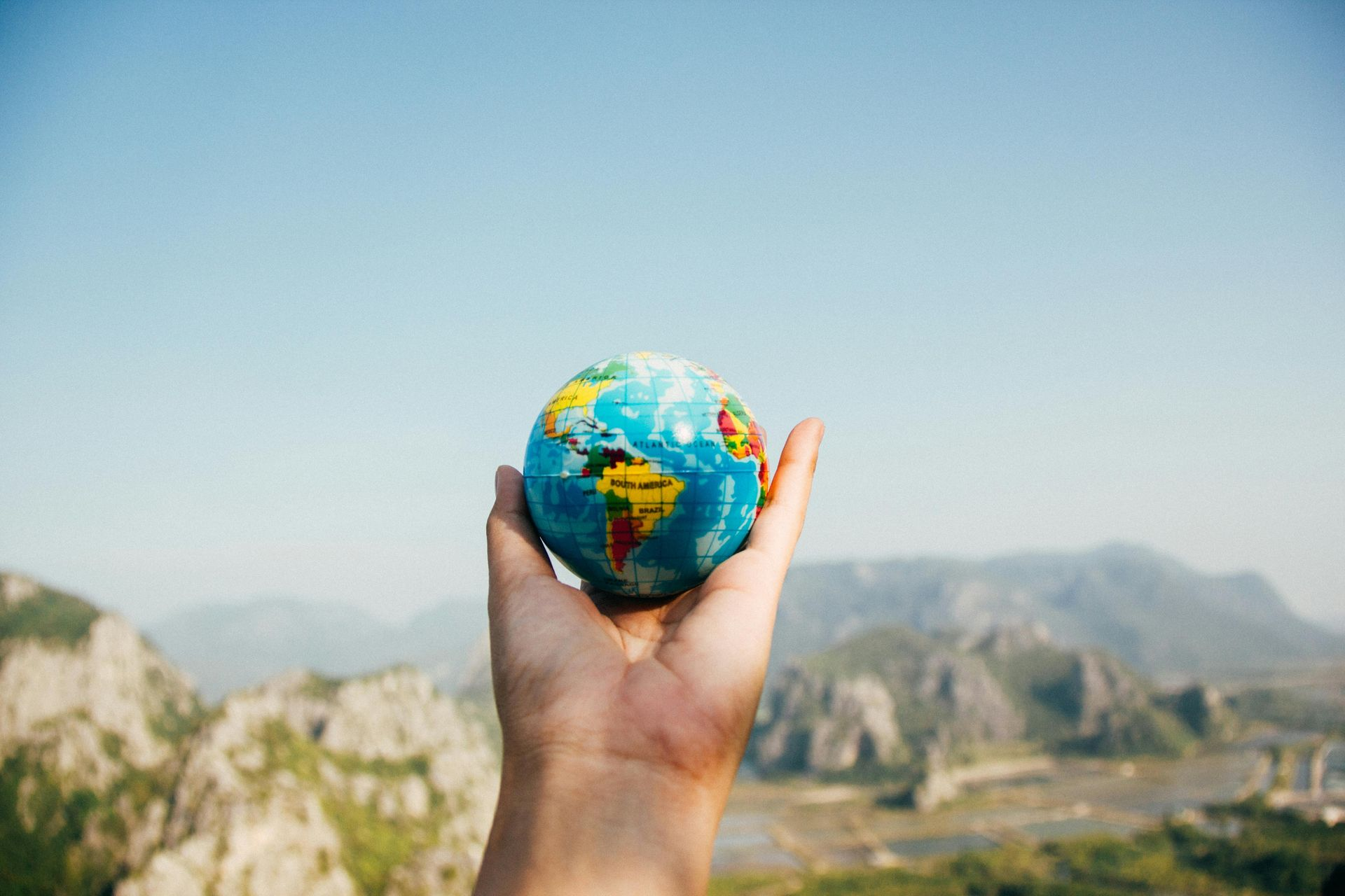 Hand holding a small globe against a backdrop of mountains and a blue sky.