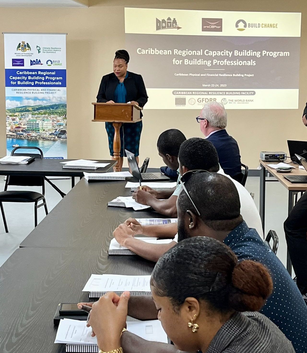 A woman is standing at a podium giving a presentation to a group of people sitting at tables.