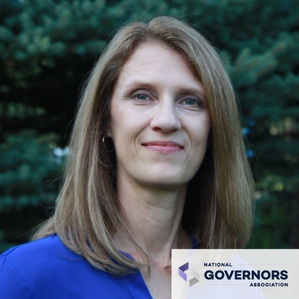 Mujer con cabello rubio hasta los hombros, sonriendo frente a la vegetación, con camisa azul. Logotipo de la Asociación Nacional de Gobernadores.