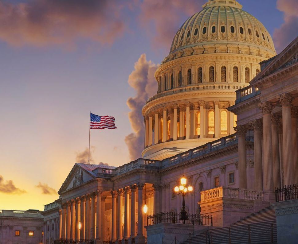 A flag is flying in front of the capitol building at sunset.