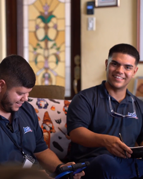 Dos hombres con camisas azules sonriendo y conversando, posiblemente en casa de un cliente. Uno sostiene un dispositivo, el otro una tableta, con una ventana decorativa al fondo.