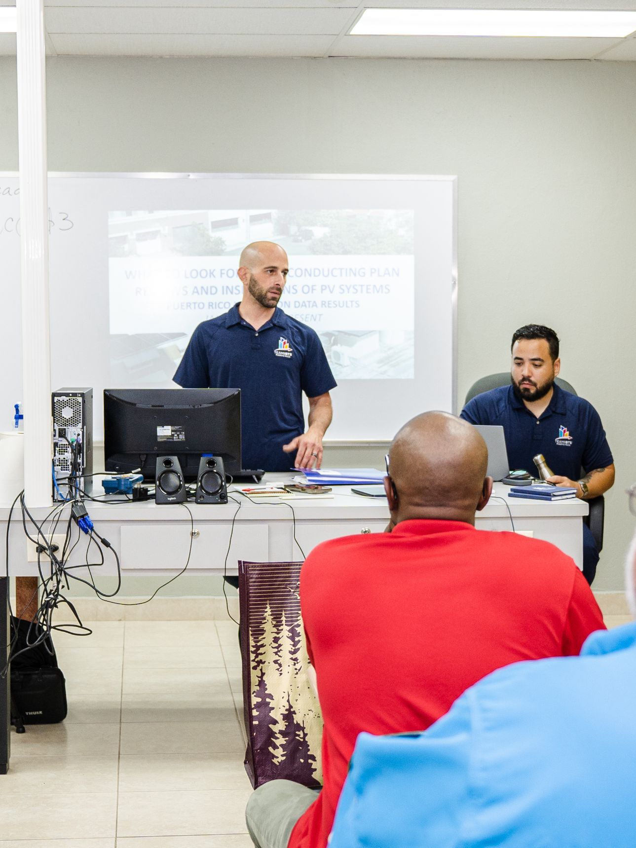 Un hombre con camisa azul presenta a un grupo pequeño. Otro hombre está sentado a una mesa con una computadora portátil, y una persona con camisa roja está en primer plano.