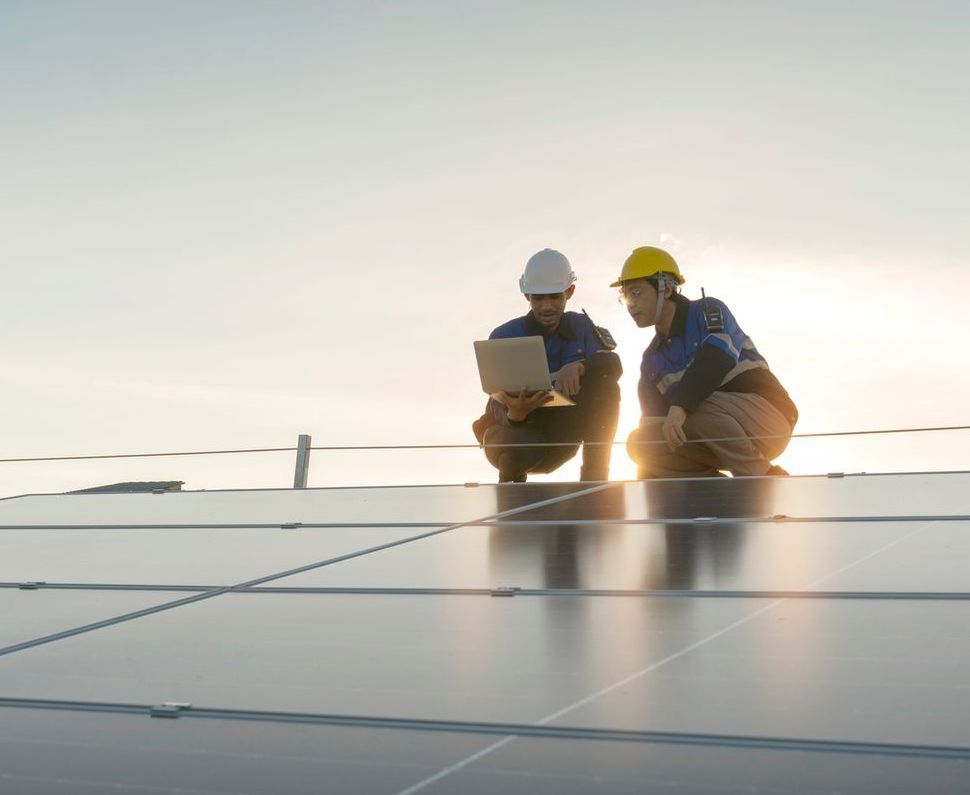 Two men are kneeling on top of a solar panel looking at a laptop.