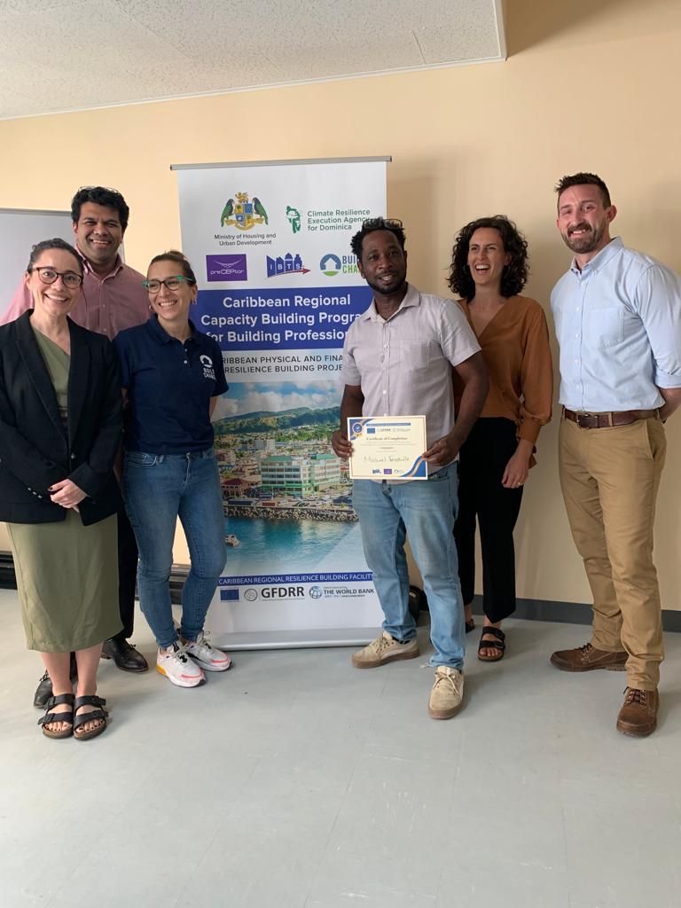 Grupo de personas frente a una pancarta; un hombre con un certificado sonríe. Dentro, probablemente una sala de trabajo o de reuniones.