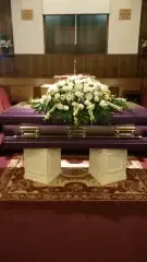 Purple casket with white floral arrangement, on two white pedestals, in a church.