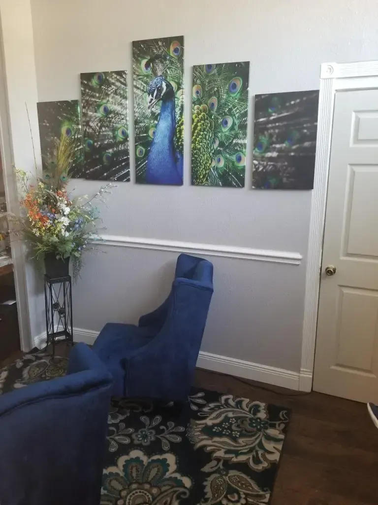 Blue peacock art on a wall above a blue chair, floral arrangement, and a patterned rug.
