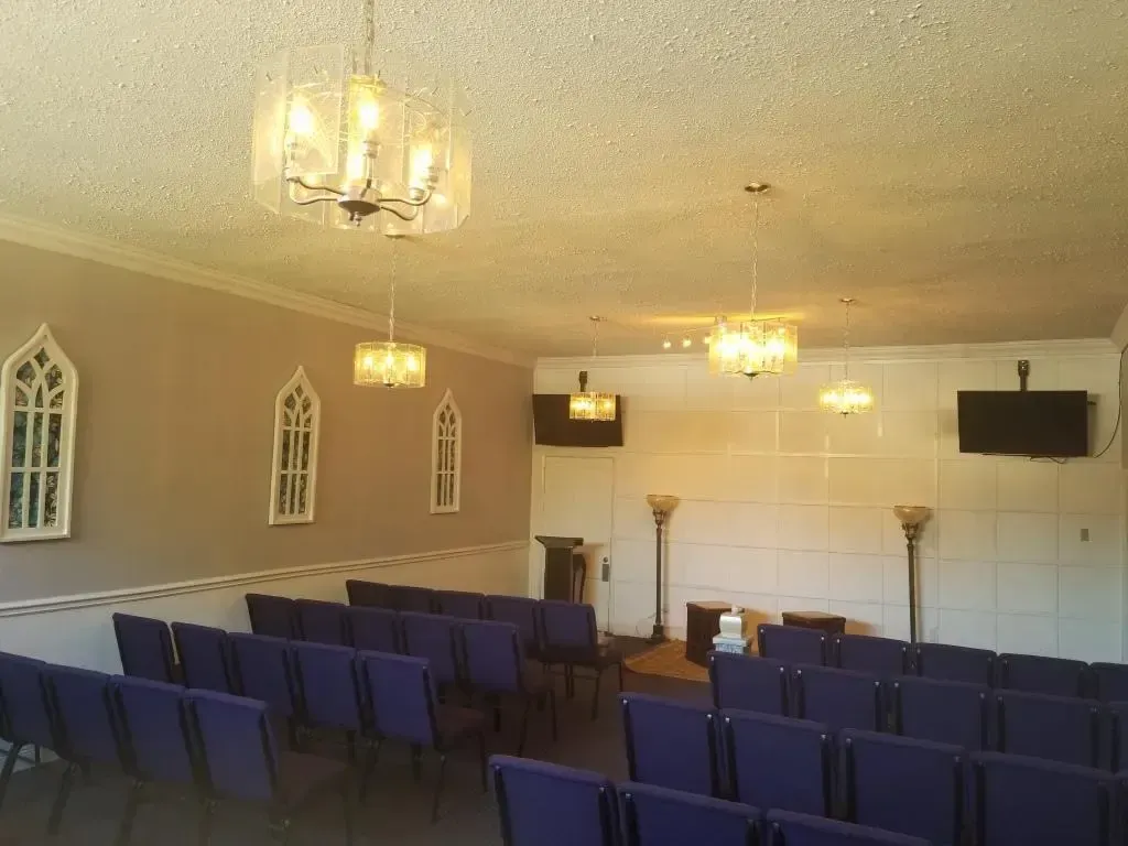 Interior of a chapel with rows of blue chairs, chandeliers, and arched windows.