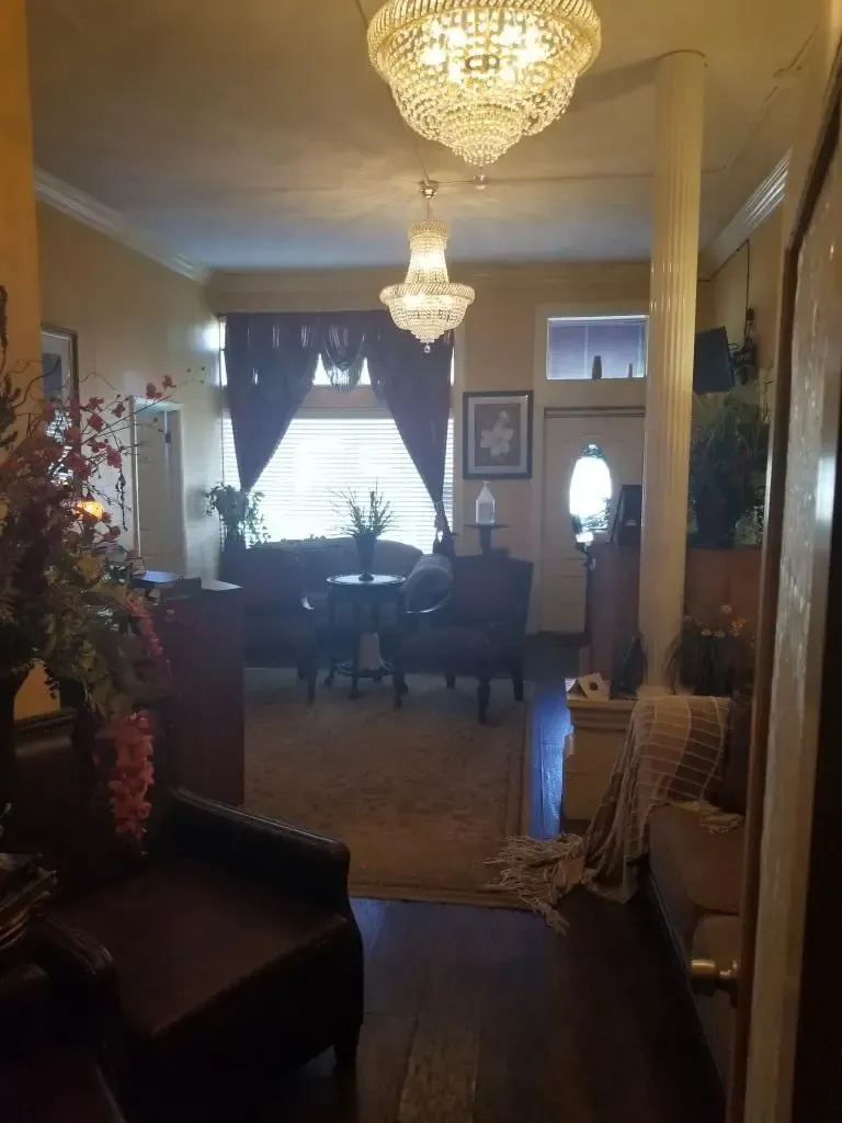 Entryway with chandeliers, dark wood floors, and a view into a living room with dark curtains and furniture.