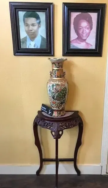 Two framed portraits above a decorative table holding a tall, ornate vase. Yellow wall.