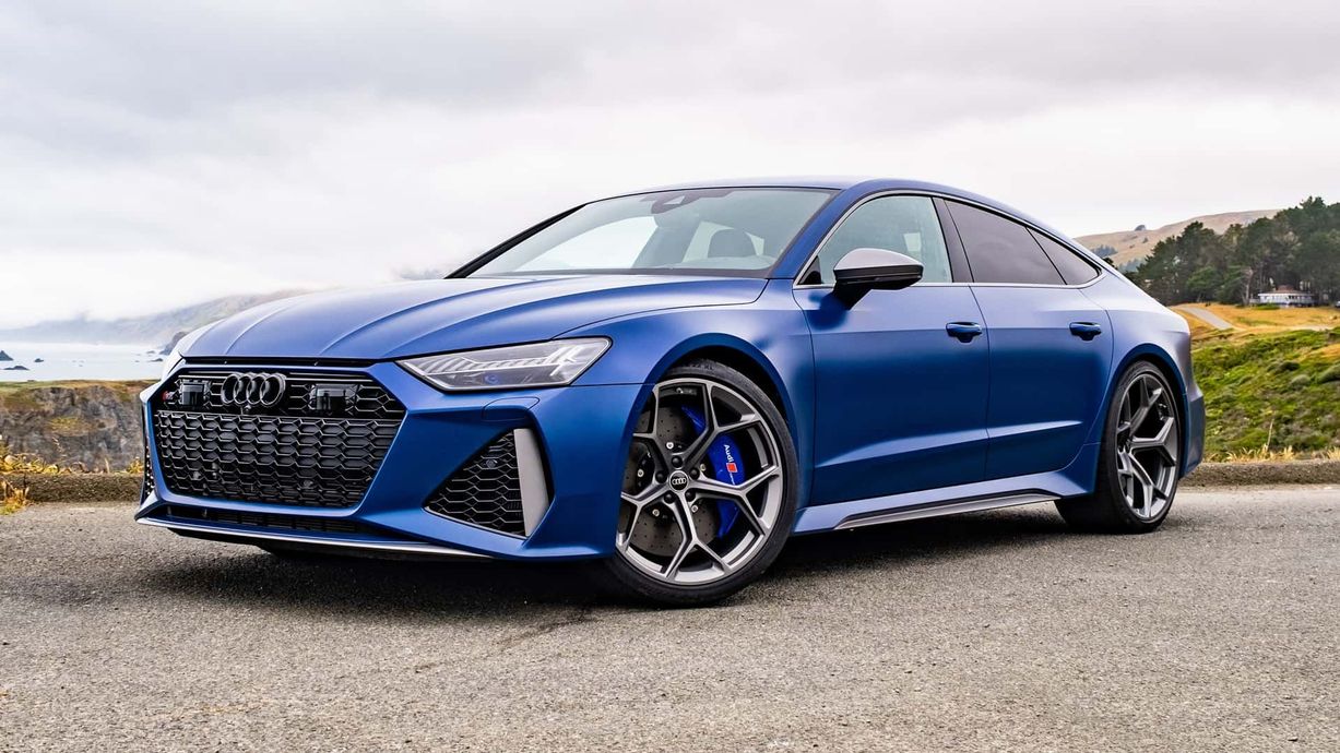 A matte blue Audi RS7 parked on a scenic coastal road overlooking the ocean.