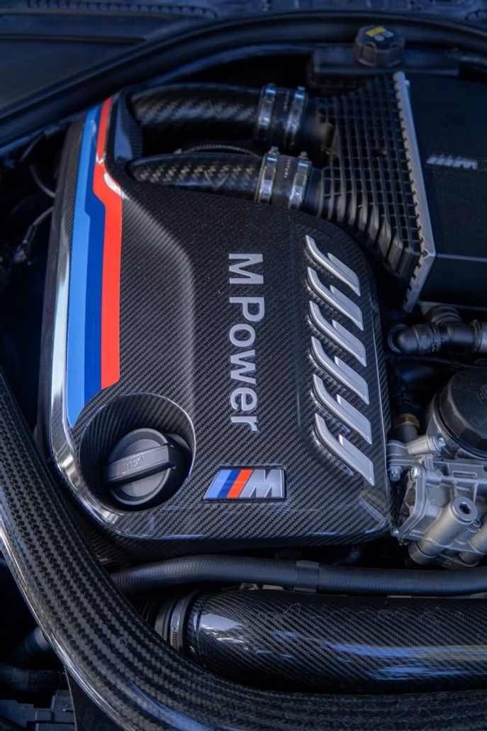 Carbon fiber BMW M Power engine cover featuring the M logo and blue, red, and light blue racing stripes.