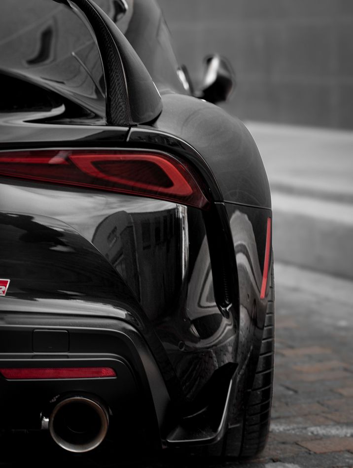 A close-up, rear-angle view of a sleek black Toyota Supra showcasing its taillight, spoiler, and exhaust pipe.