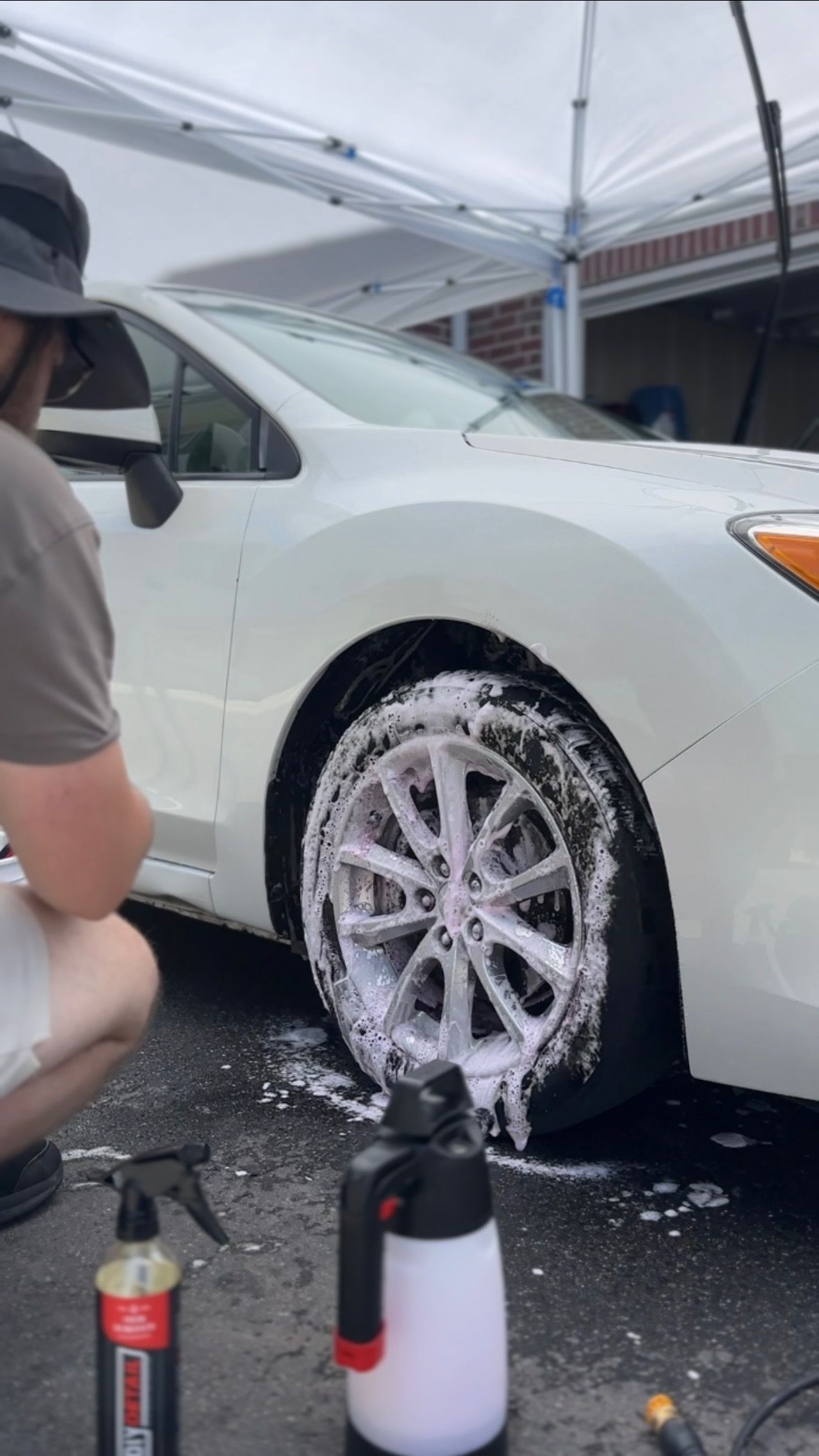 Person washing a white car tire with foam. Outdoors, under a canopy.