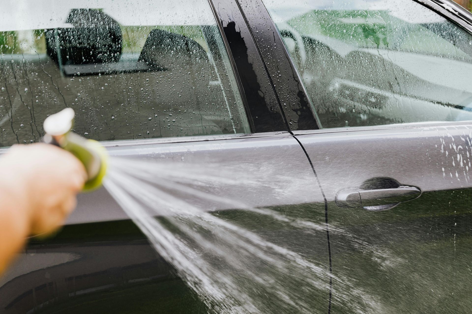Person spraying water on a dark car with a hose in an outdoor setting.