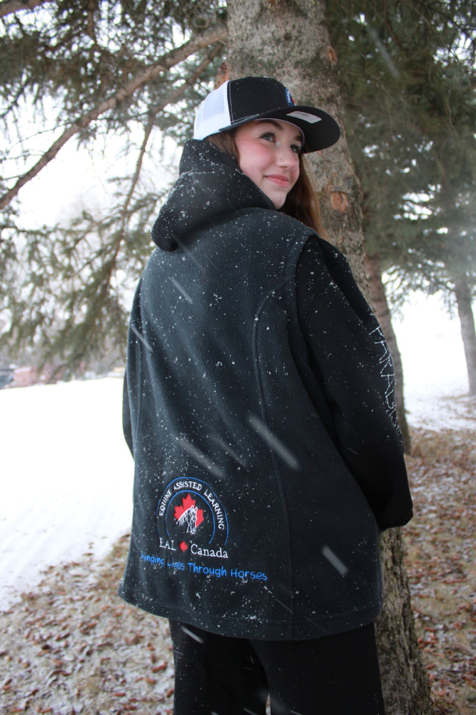 Woman in black hat and hoodie smiles next to a tree in a snowy outdoor setting.